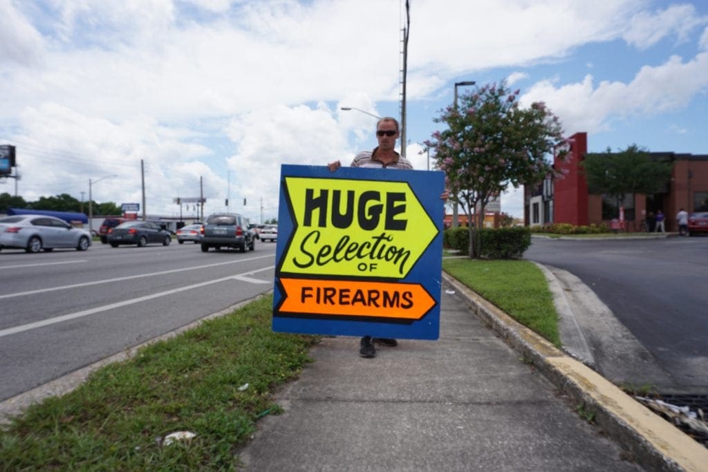 Man holding sign with arrow pointing that reads Huge Selection of Firearms. 